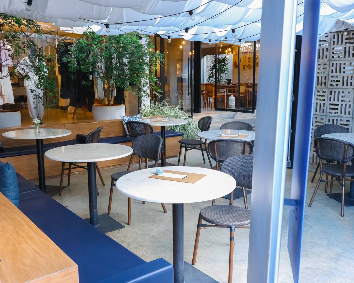 Ambiente externo da Casa Belgaro, com mesas redondas de mármore, banco estofado azul, plantas e tecidos claros no teto