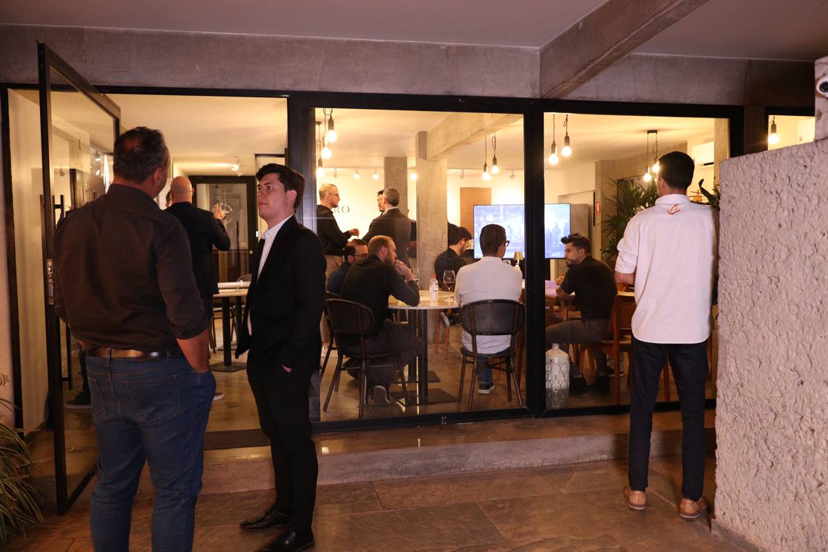 Fachada de vidro da Casa Belgaro à noite, com o salão iluminado e cheio
