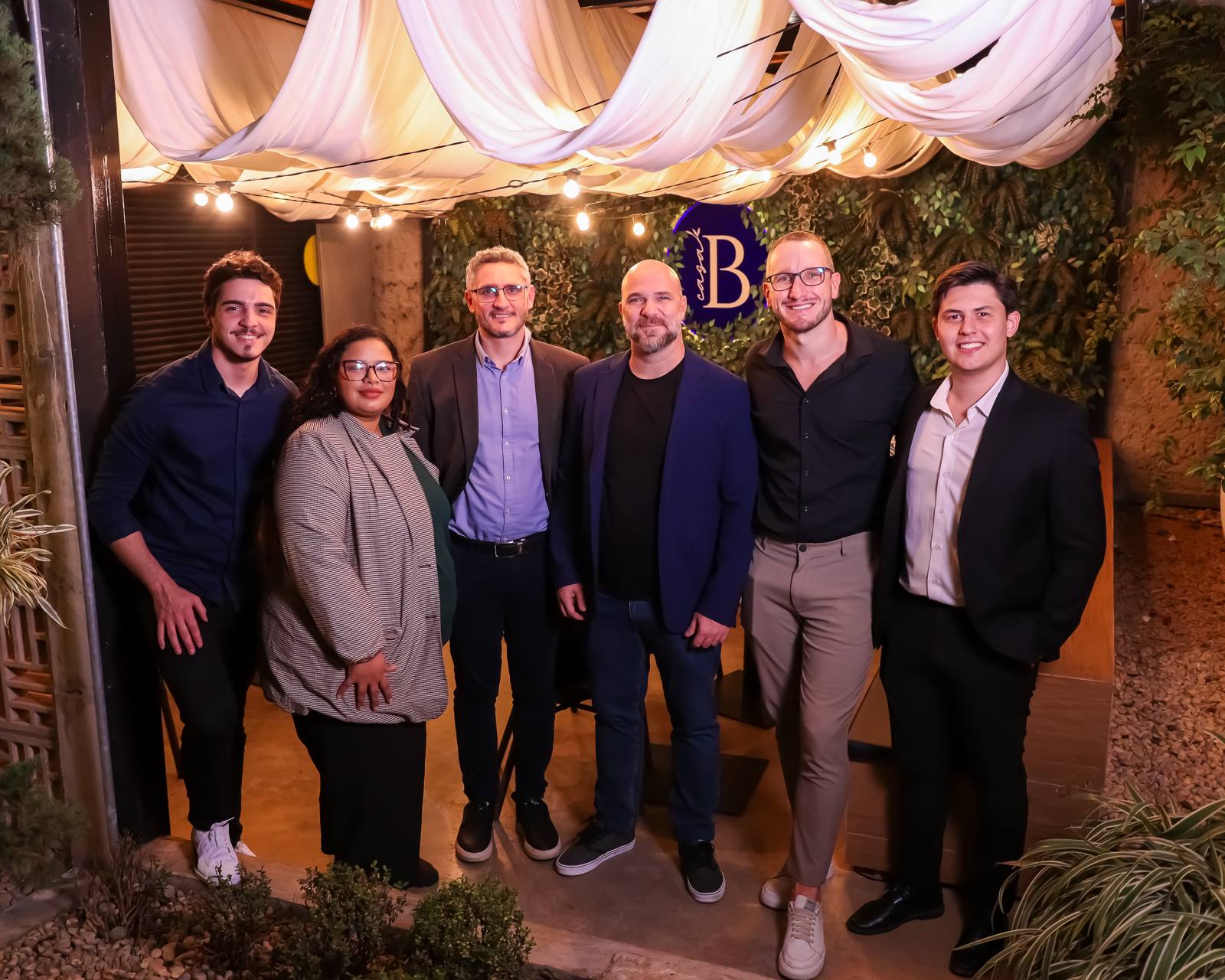 Ambiente externo da Casa Belgaro à noite, com tecidos, varal de luzes e o painel verde com o B da casa